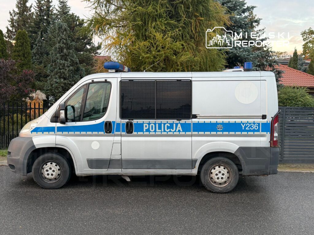 Funkcjonariusz policji w radiowozie parkowanym na ulicy, w tle ogrodzenie i drzewa.