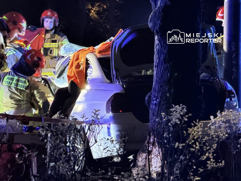 Grupa strażaków w hełmach pracuje przy rozbitym samochodzie, otwierając bagażnik w ciemnej okolicy.