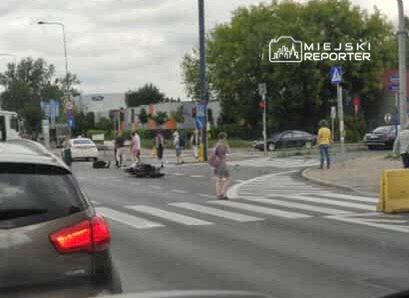 Wypadek drogowy na przejściu dla pieszych. Na jezdni leży motocykl, a obok zatrzymali się świadkowie zdarzenia. W tle widać samochody i przechodniów. Sytuacja wymaga interwencji służb ratunkowych. Zdarzenie miało miejsce w mieście, w okolicy ruchliwego skrzyżowania.