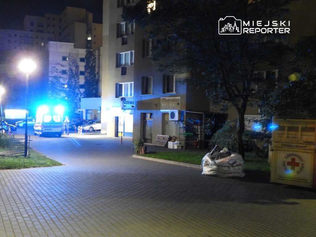 Karetka z włączonymi sygnałami świetlnymi stoi na osiedlowej ulicy w nocy, obok budynku mieszkalnego.