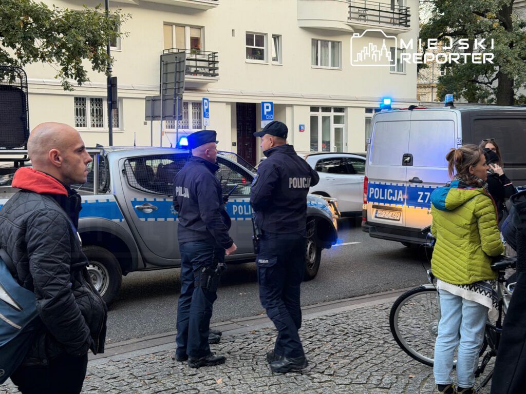 Policjanci w mundurach rozmawiają obok radiowozu z włączonymi sygnałami świetlnymi na miejskiej ulicy.