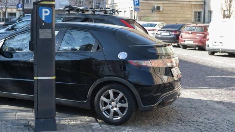 Czarny samochód zaparkowany obok parkomatu na ulicy, w tle widoczne inne pojazdy oraz budynki.