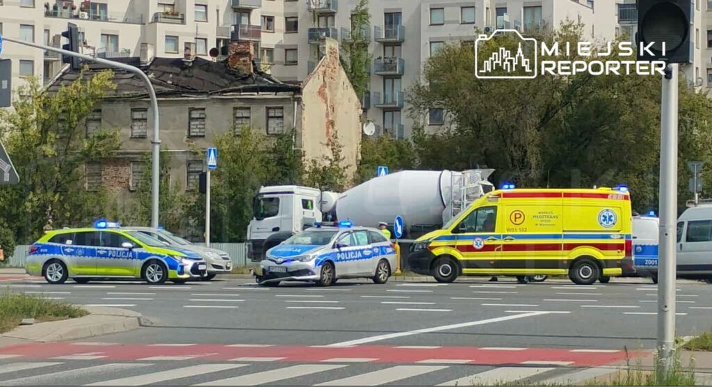 Policyjny radiowóz z włączonymi sygnałami świetlnymi blokuje ruch na skrzyżowaniu obok karetki pogotowia i betoniarki.