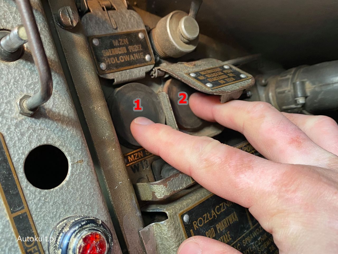 Naciśnij najpierw przycisk (1) bez klapki (słychać mechanizm pompy oleju), a następnie, trzymając go, wciśnij przycisk (2) pod klapką – to przycisk rozrusznika. Trzymaj, aż silnik się uruchomi