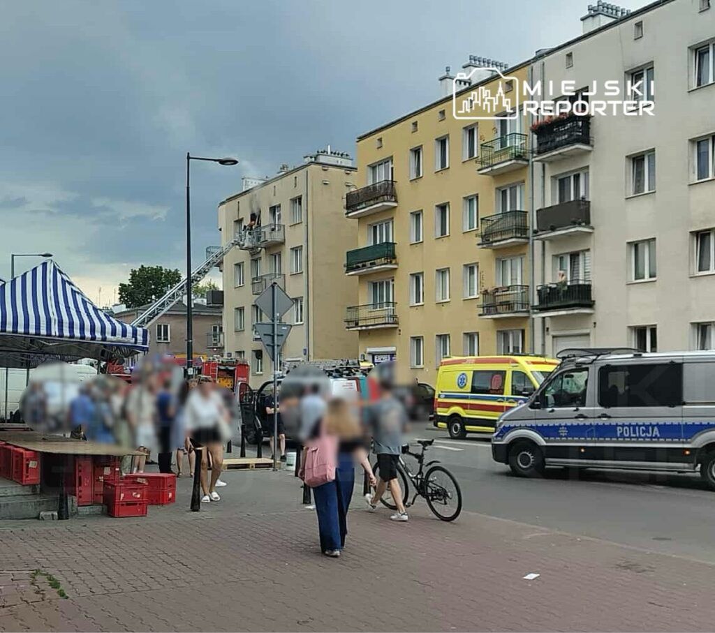 Grupa ludzi gromadzi się przy straganie, podczas gdy straż pożarna i karetka stoją na ulicy obok budynku mieszkalnego.