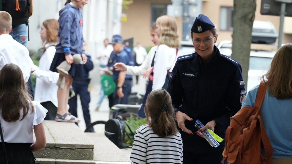 Policjantka rozmawia z dzieckiem na chodniku, w tle widać grupę uczniów i mundurowych.
