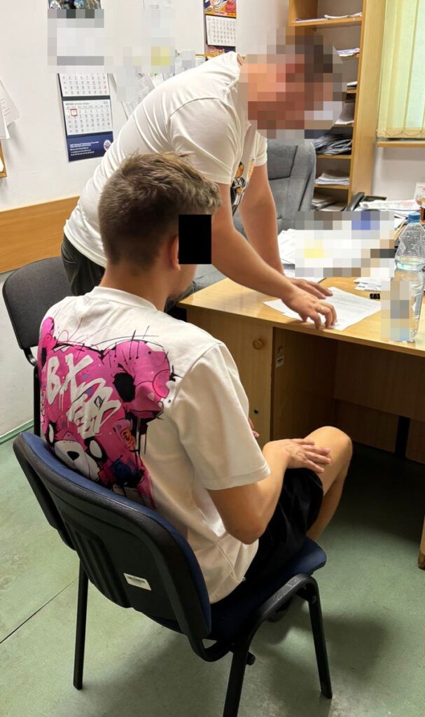 Mężczyzna w białej koszulce z kolorowym nadrukiem siedzi na krześle, podczas gdy drugi mężczyzna pochyla się nad biurkiem w biurze.