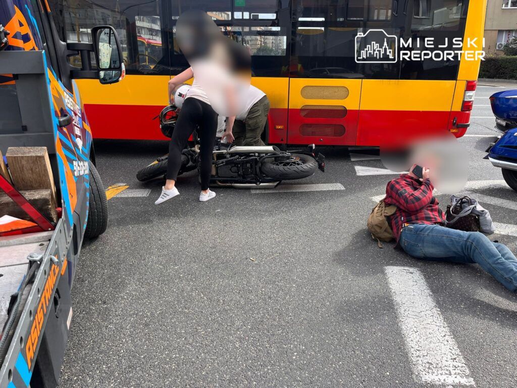 Wypadek drogowy z udziałem motocykla i autobusu miejskiego. Na miejscu zdarzenia osoby próbują pomóc poszkodowanemu motocykliście, który leży na jezdni. Widoczny również kierowca autobusu. W tle widać inne pojazdy. Służby ratunkowe mogą być w drodze.