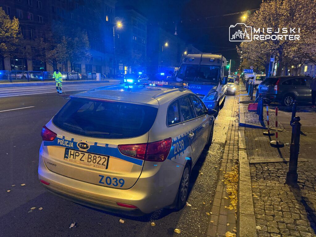 Policyjna Kia zaparkowana na ulicy, z włączonymi sygnałami świetlnymi, obok stoi radiowóz. W tle widać oświetloną ulicę.