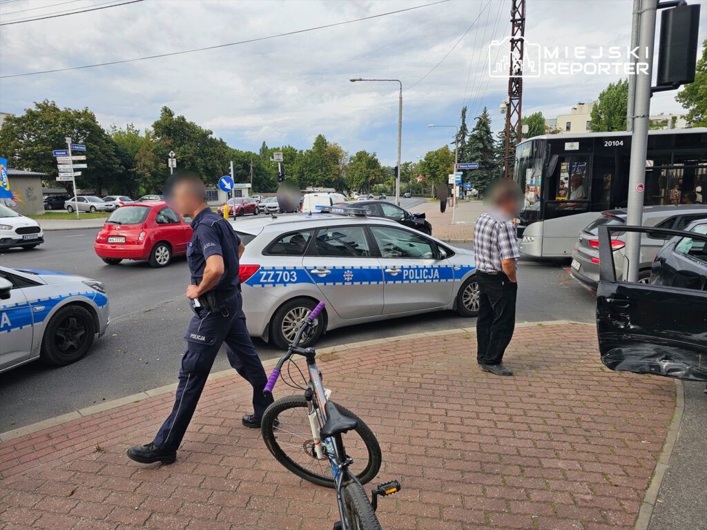 Policjant w mundurze przechodzi obok uszkodzonego pojazdu na skrzyżowaniu, a mężczyzna w koszuli obserwuje sytuację.