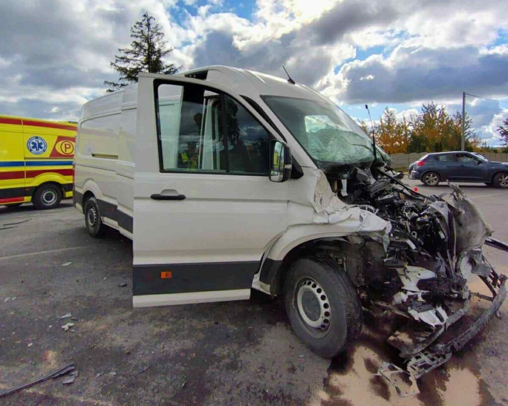 Rozbity biały furgon z uszkodzoną przednią częścią stoi na parkingu, obok widać ambulans.
