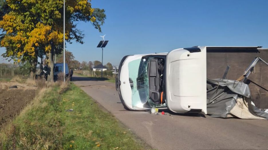 Przewrócony samochód dostawczy leży na boku na drodze, w otoczeniu drzew i pól.