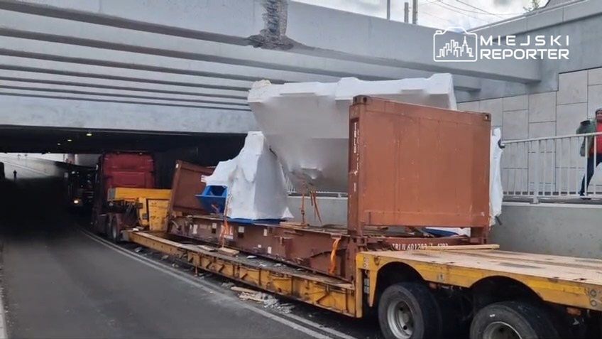 Ciężarówki przewożące ładunek z folią ochronną przejeżdżają pod niskim wiaduktem drogowym.