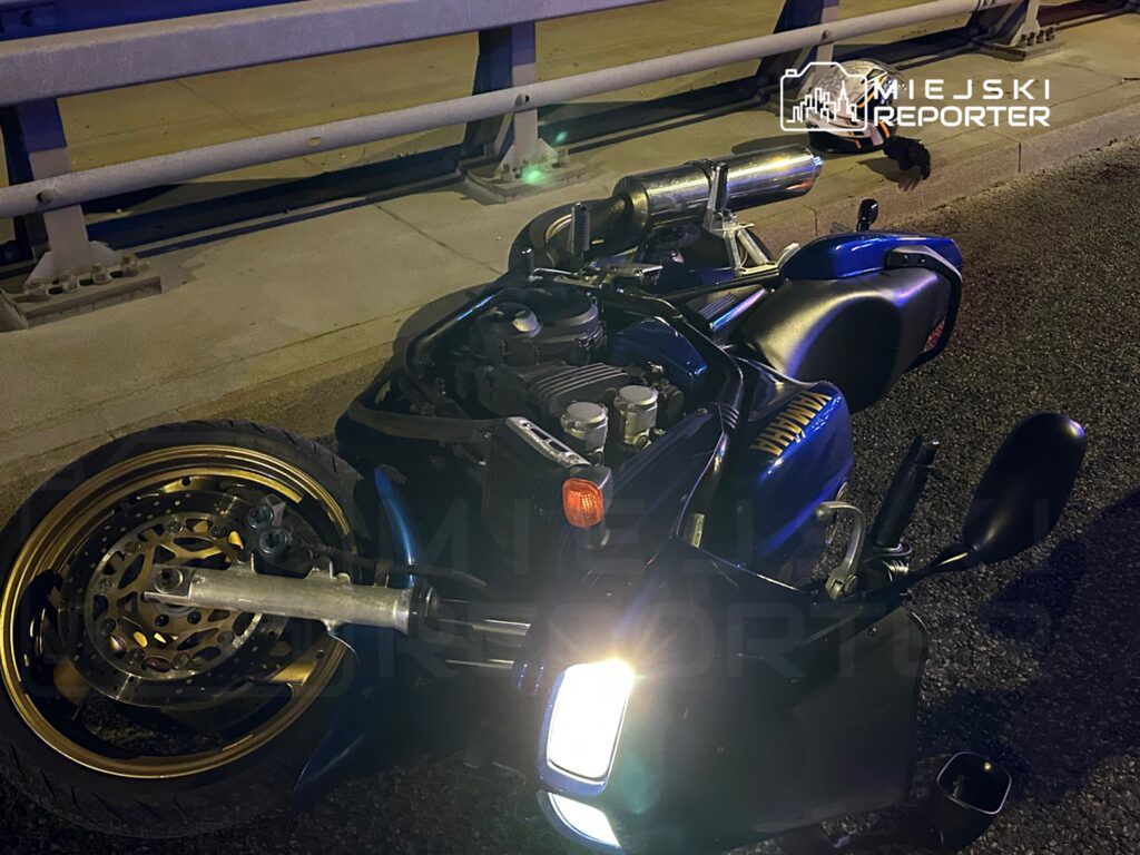 Motocykl leżący na boku na jezdni, z widocznymi uszkodzeniami oraz odblaskowym światłem.