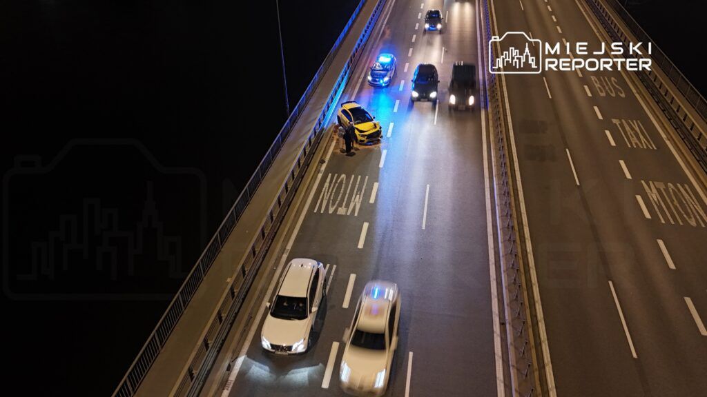 Na moście zatrzymany jest żółty samochód, obok stoją policjanci, a w tle widać pojazdy z włączonymi sygnałami świetlnymi.