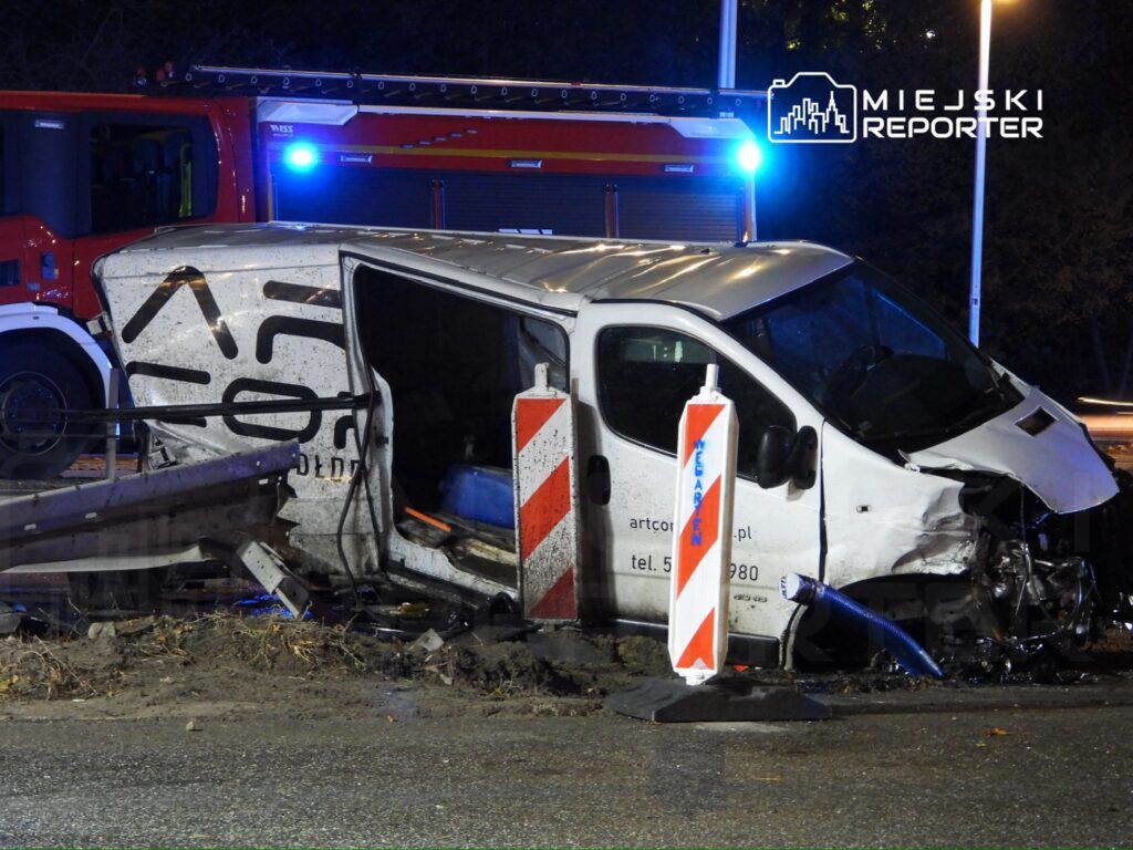 Rozbity samochód dostawczy z uszkodzoną boczną częścią stoi przy barierkach na drodze, obok wozu strażackiego.