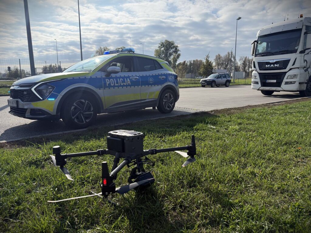 Dron z kamerą znajduje się na trawie obok radiowozu policji, a w tle przejeżdża ciężarówka.