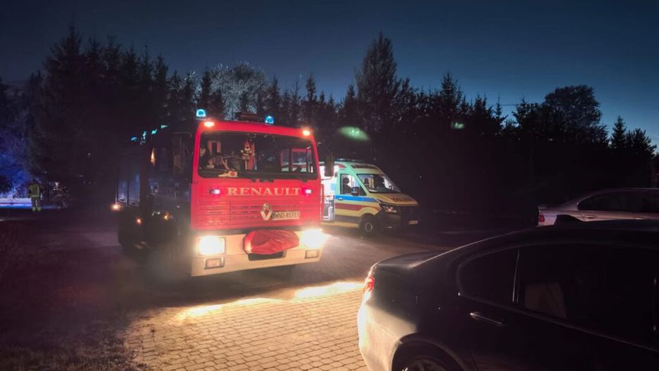 Czerwony wóz strażacki z niebieskimi światłami parkowany na drodze, obok karetka pogotowia w nocy.