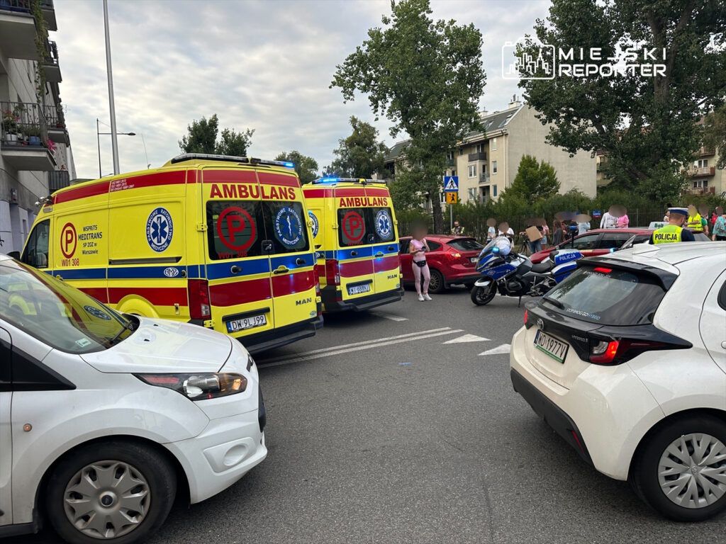 Dwa karetki pogotowia stoją na ulicy, obok policyjny motocykl, a w tle grupa ludzi obserwujących zdarzenie.