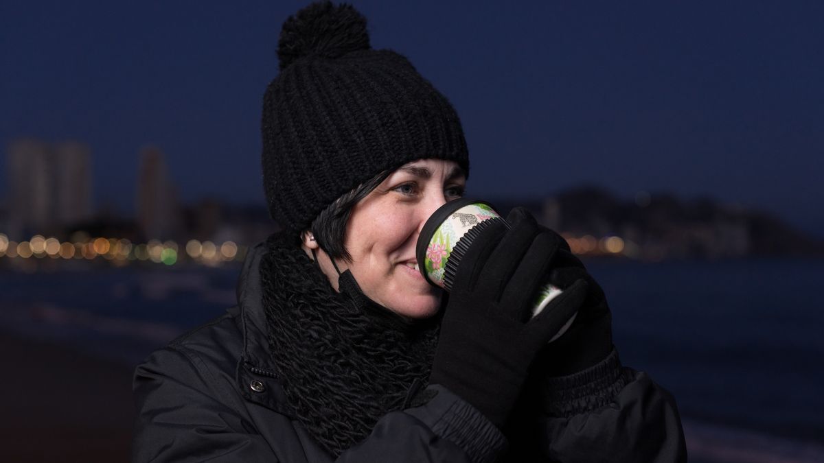 Young woman smiling on a very cold day and drinking hot drink on the beach at night with the city lights in the background.
Young woman smiling on a very cold day and drinking hot coffee on the beach at night with the city lights in the background. She wears black clothes and a black hat, gloves and takes off her protective face mask to drink.
machirito
