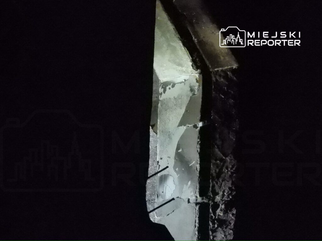 Zniszczona ściana budynku z widocznymi fragmentami tynku i oświetleniem w tle.