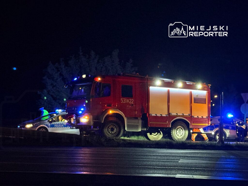 Straż pożarna i policja interweniują na drodze w nocy, przy użyciu sygnalizacji świetlnej.