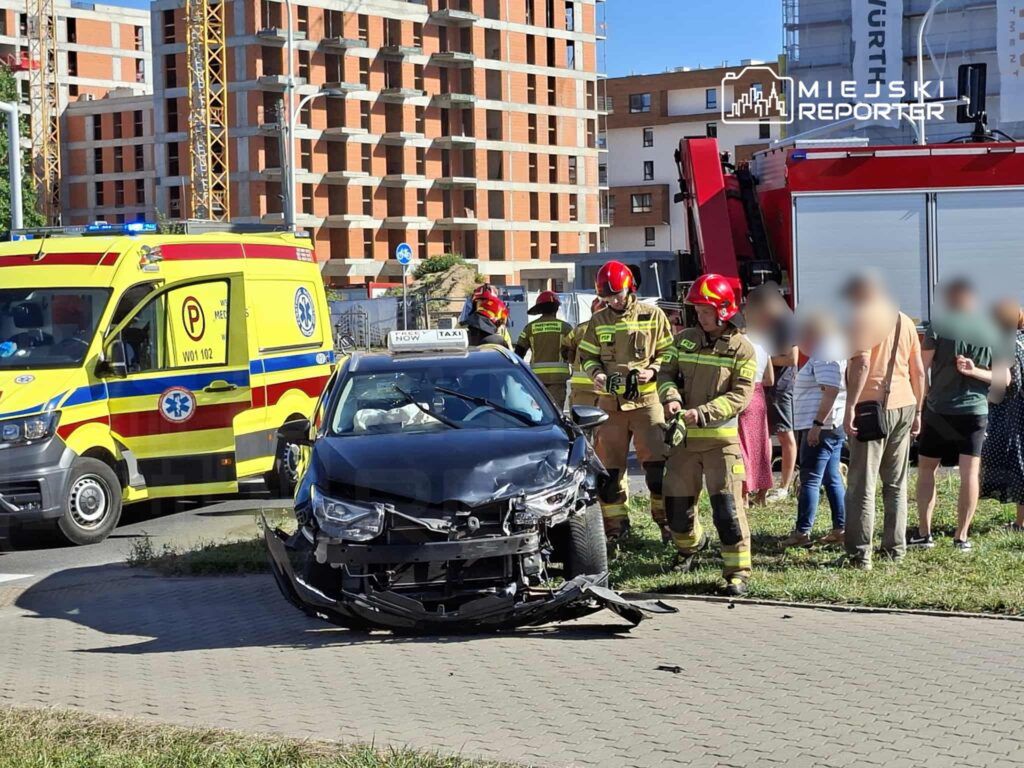 Czarny samochód osobowy z uszkodzoną przednią częścią, obok strażacy w hełmach oceniają sytuację przy krawężniku.