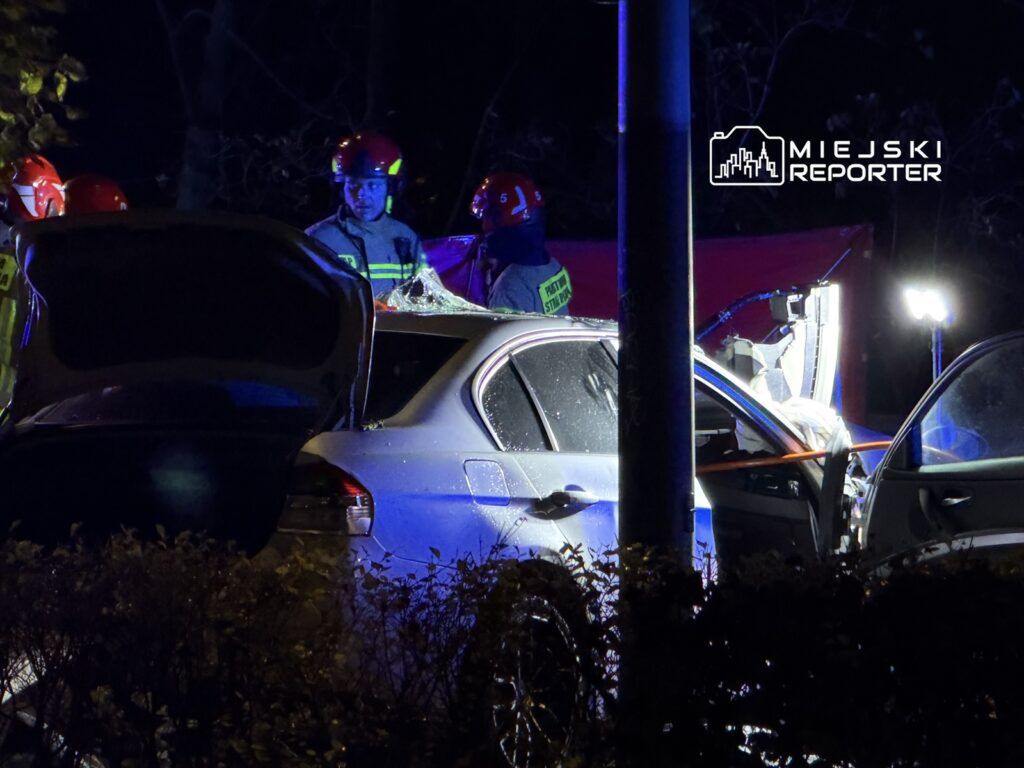 Grupa strażaków w hełmach pracuje przy uszkodzonym samochodzie w nocy, oświetlonym przez lampy ratunkowe.