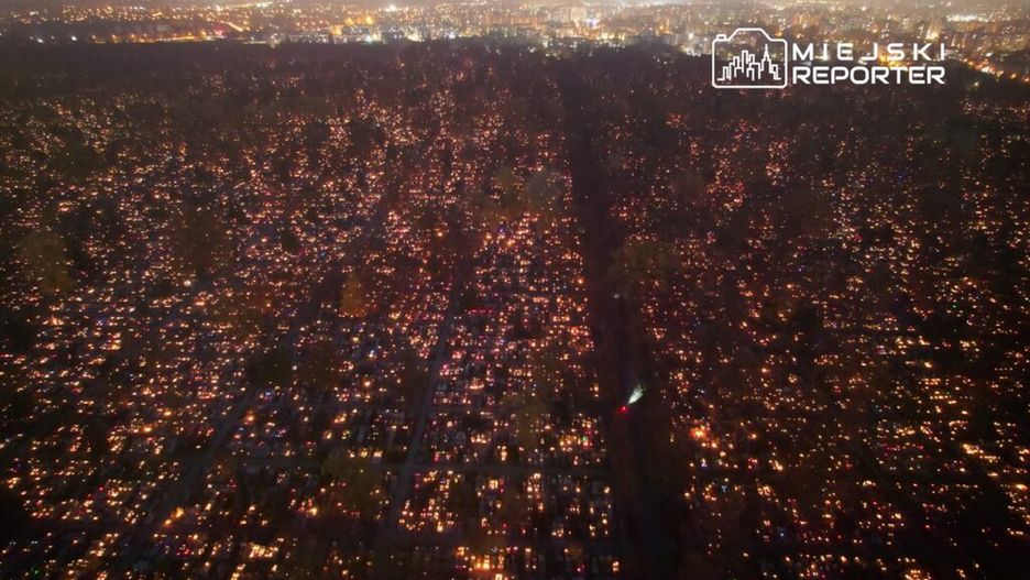 Widok z lotu ptaka na cmentarz, w którym setki zniczy oświetlają groby w ciemności, tworząc świetlną mozaikę.