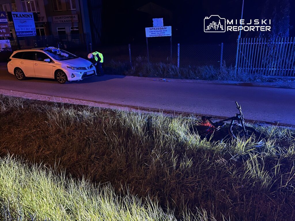 Policjant w odblaskowej kamizelce bada uszkodzony samochód osobowy obok leżącego roweru na poboczu drogi.