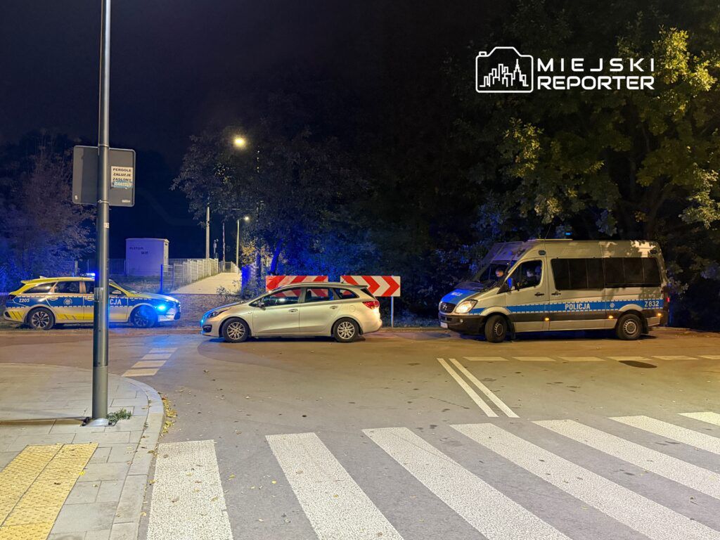 Policyjny radiowóz oraz van stoją na ulicy, blokując ruch, w otoczeniu oświetlonym przez latarnie.