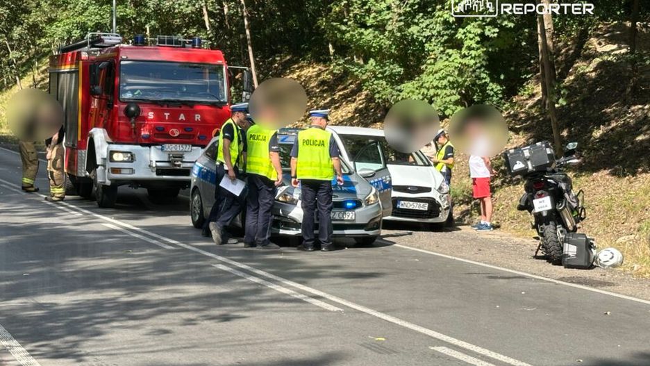 Grupa policjantów rozmawia z kierowcą uszkodzonego samochodu na poboczu drogi, obok stoi wóz strażacki.