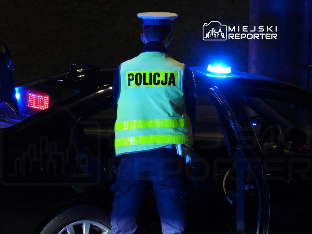 Policjant w odblaskowej kamizelce kontroluje samochód przy zapalonych sygnałach świetlnych.