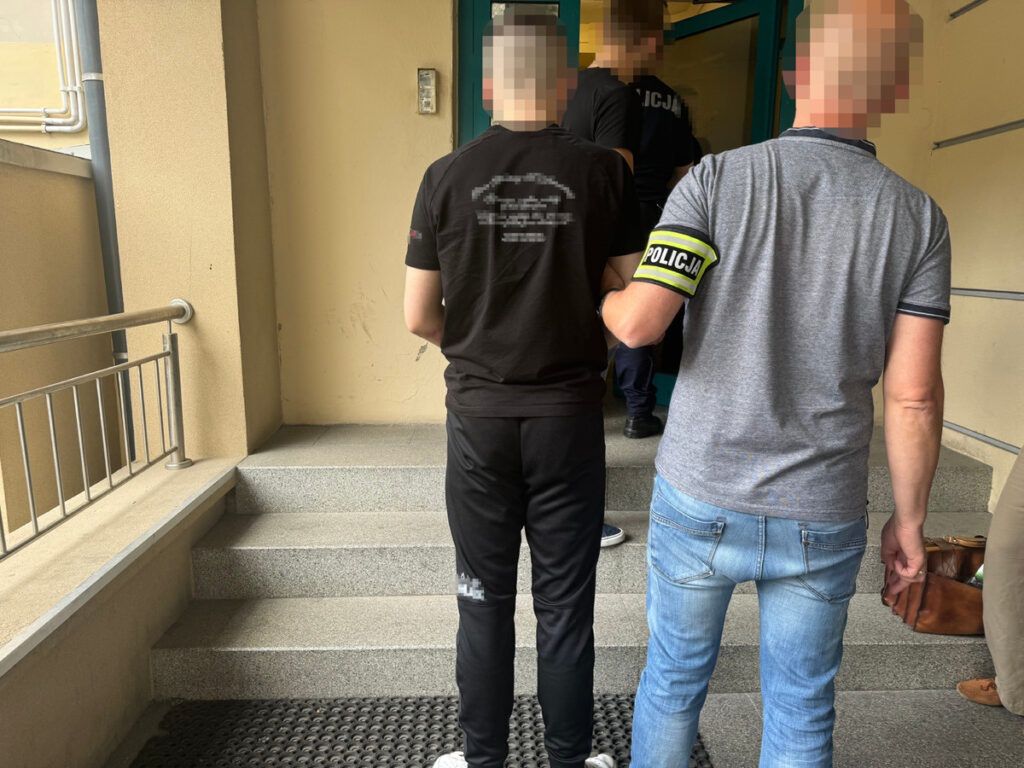 Mężczyzna w czarnej koszulce prowadzony przez funkcjonariusza policji na schodach budynku.