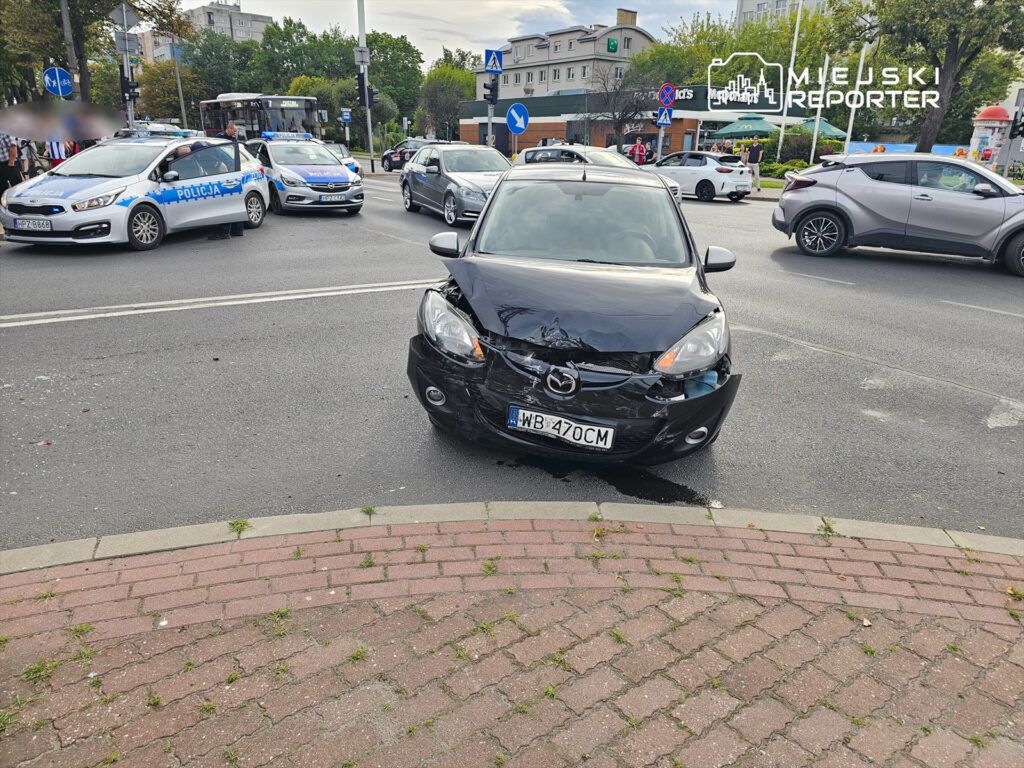 Czarny samochód osobowy z uszkodzoną przednią częścią stoi na skrzyżowaniu, otoczony przez radiowozy policji.