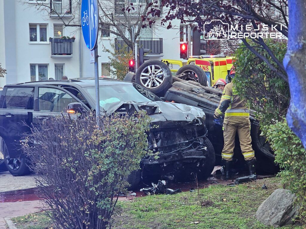 Zniszczony samochód leży na boku obok drogi, a strażak pomaga osobie w środku pojazdu. W tle widać sygnalizację świetlną i karetki.