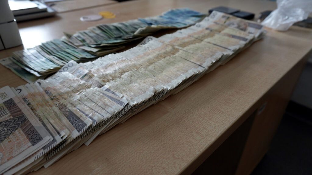 Stos równo ułożonych banknotów w walucie krajowej na drewnianym biurku, obok leżą inne banknoty oraz telefon komórkowy.
