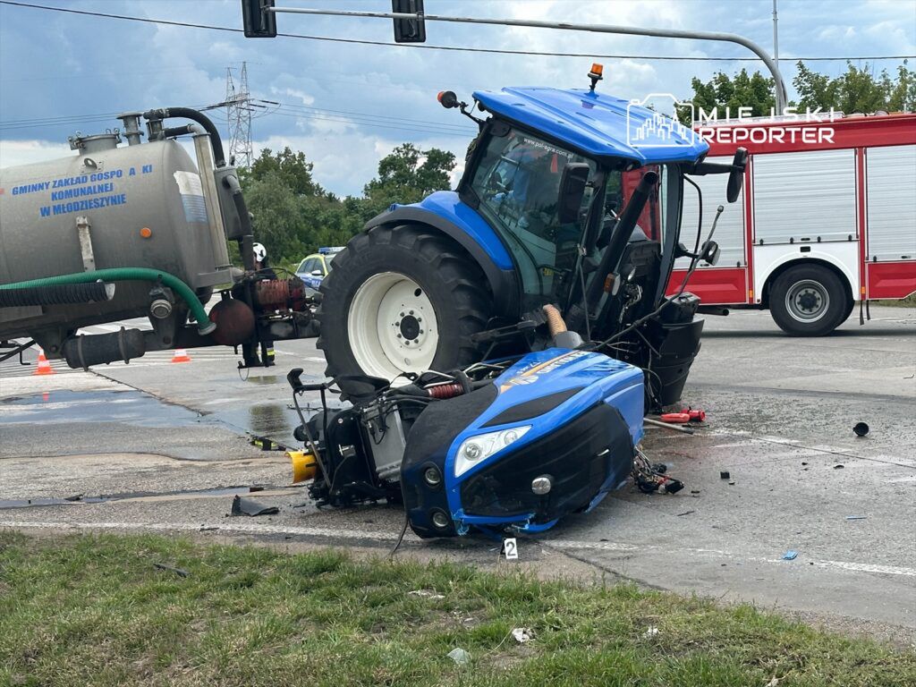 Traktor przewrócony na drodze, obok leży uszkodzona maszyna, a w tle widoczny wóz strażacki.
