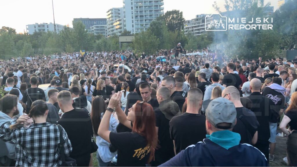 Tłum ludzi bawiący się na otwartym terenie, w tle nowoczesne budynki oraz zieleń drzew.