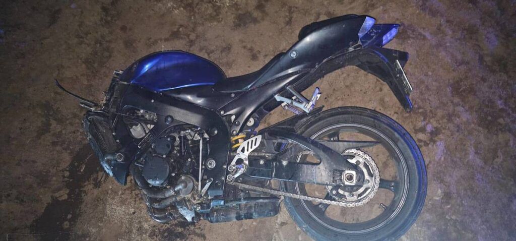 Uszkodzony czarny motocykl leży na ziemi w nieoświetlonym otoczeniu, z widocznymi zarysowaniami i zabrudzeniami.