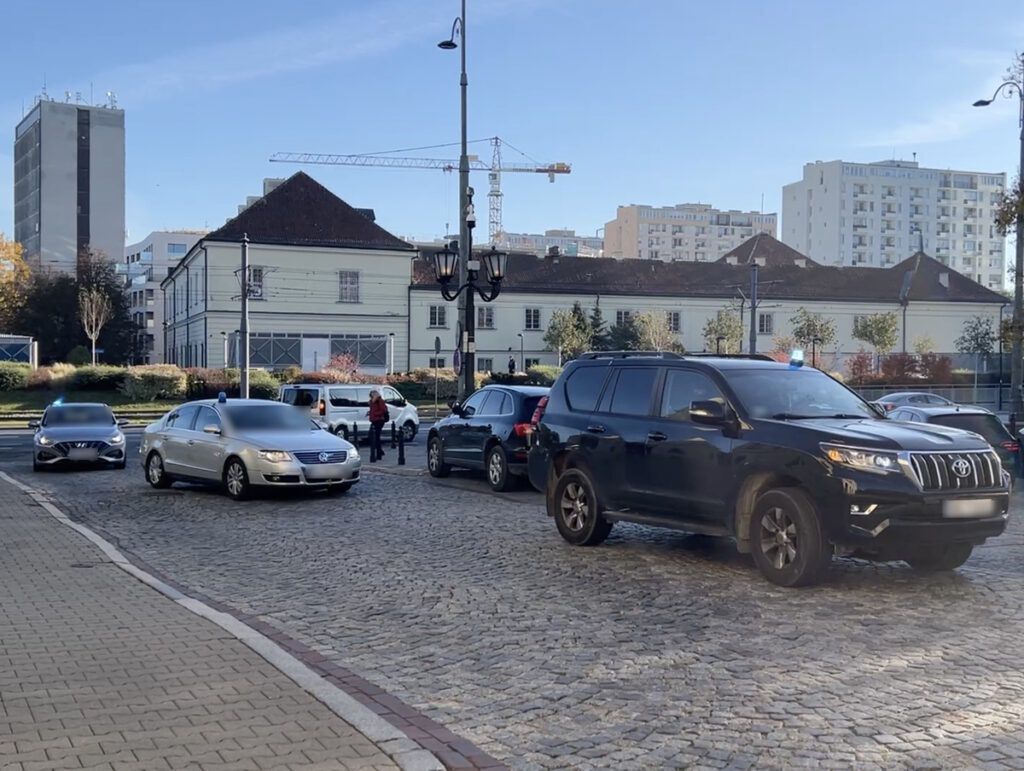 Ruchome pojazdy na brukowanej ulicy, w tle nowoczesne budynki i plac z drzewami.