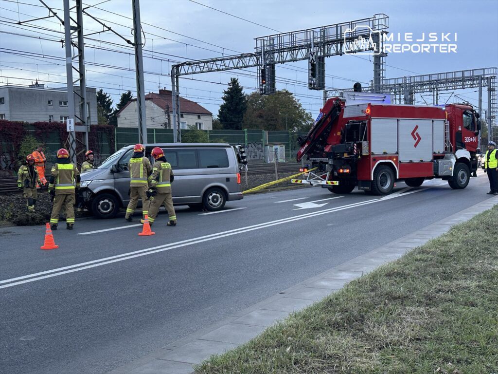 Grupa strażaków w hełmach pracuje przy uszkodzonym pojeździe, obok stoi wóz strażacki z podnośnikiem.