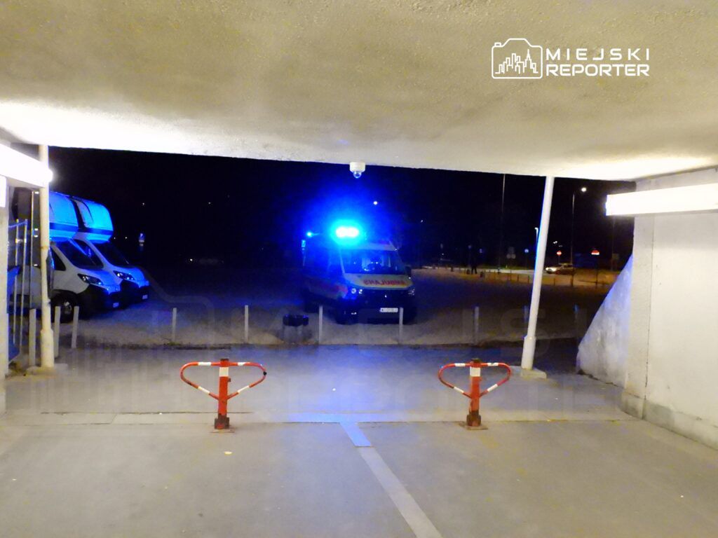 Karetka pogotowia z włączonymi sygnałami świetlnymi stoi na parkingu nocą, w tle widoczne inne pojazdy.
