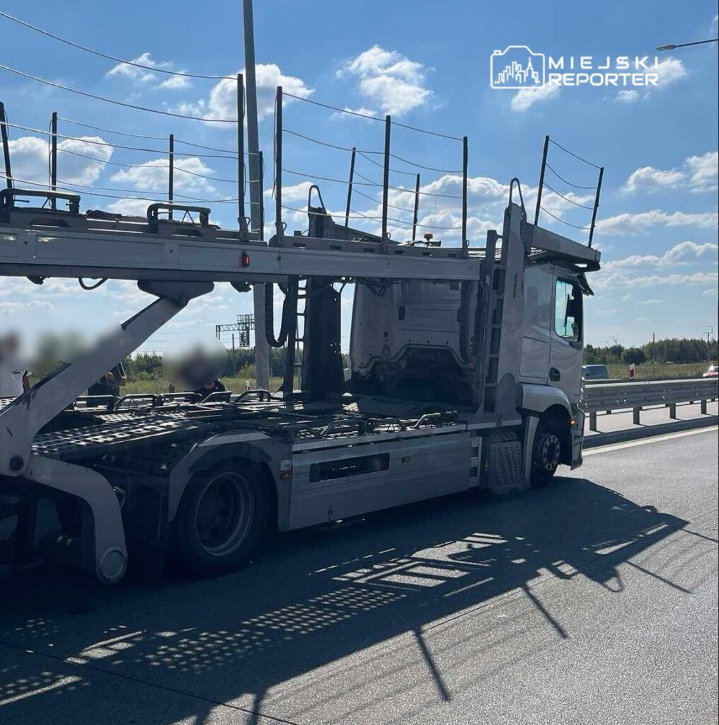 Ciężarówka do transportu samochodów zatrzymana na pasie drogowym w słoneczny dzień, z widocznymi chmurami na niebie.