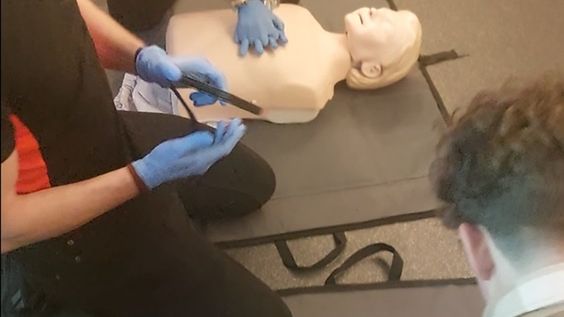 Mężczyzna w rękawiczkach wykonuje resuscytację na manekinie, podczas gdy kobieta obserwuje z boku.