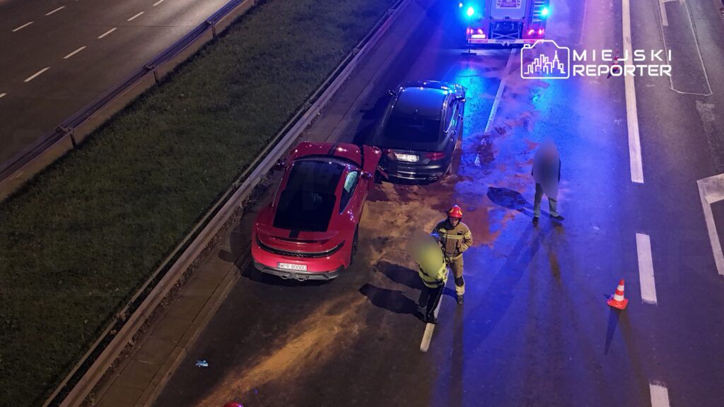 Na drodze stoi czerwony sportowy samochód, zderzony z czarnym pojazdem. Strażak rozmawia z policjantem, w tle widać włączone sygnały świetlne.
