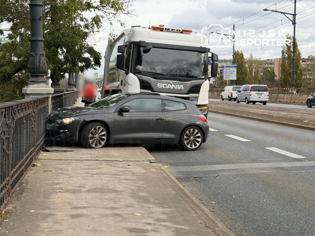 Czarny samochód osobowy stoi na chodniku, zderzony z barierką, obok niego przejeżdża ciężarówka.