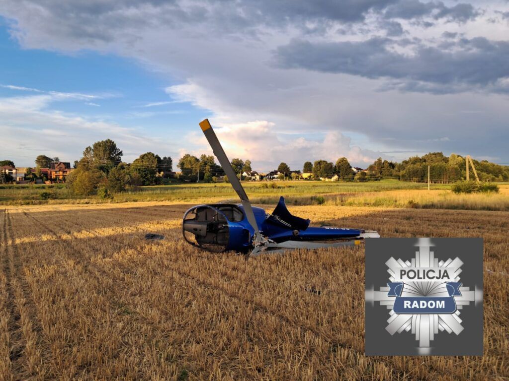 Helikopter leży na polu z zebranym zbożem, w tle widoczne domy oraz drzewa.