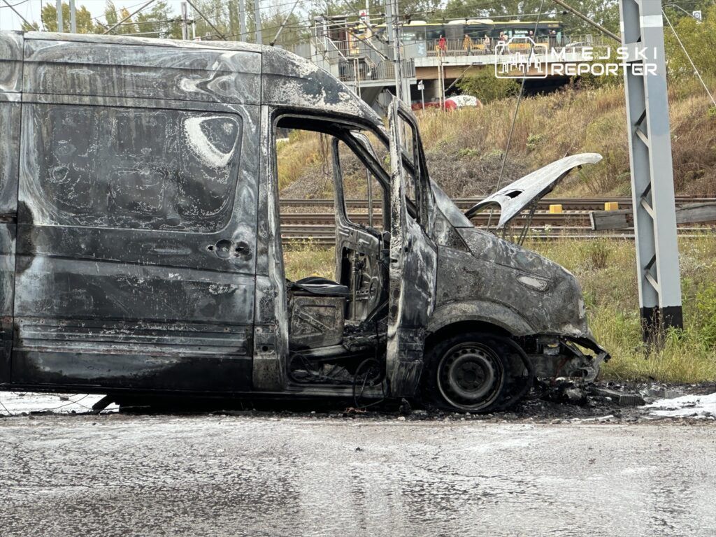 Spalony furgon stojący na poboczu drogi, z widocznymi uszkodzeniami karoserii i zniszczonym wnętrzem.