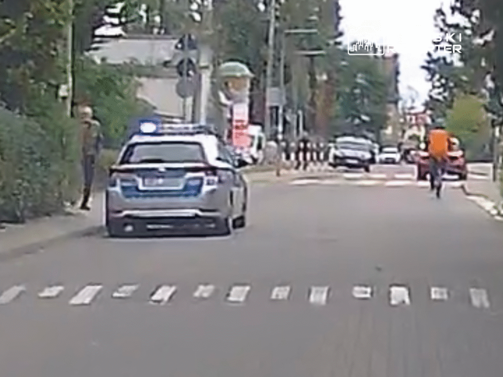 Radiowóz policyjny zatrzymany przy krawężniku, a w tle rowerzysta w pomarańczowej koszulce przejeżdża obok.